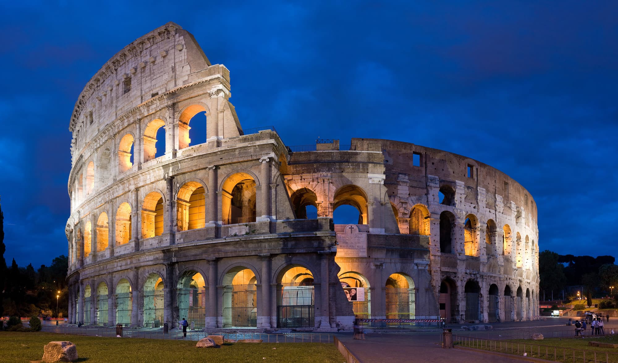 The Colosseum in Rome lit at dusk, with arches and exterior walls visible.